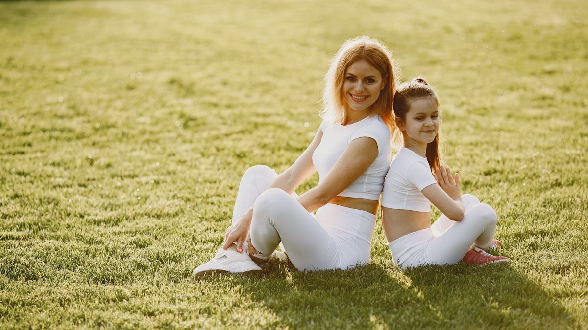 A mother and daughter sit back to back in matching activewear on a sunny grass field, enjoying a summer day together.